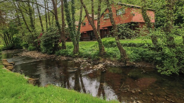 A red cabin with a river in front, surrounded by trees and greenery. - Cabin No. 12 - Salt and Fern Cabins (Port Talbot)