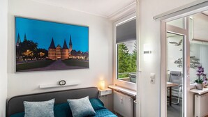 A hotel room with a bed, a large window, a radiator, a small table, and a wall-mounted picture. - Apartment 'Lubeca' with Loggia (Goslar)