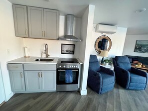 A compact kitchen with grey cabinets, a stainless steel range, and a built-in oven. There are two blue-striped armchairs and a small side table with a lamp. - Simply Saugy - A Relaxing Saugatuck Getaway! (Saugatuck)