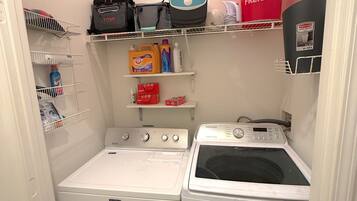 Een wasruimte met een Maytag wasmachine en een Samsung droger, planken met schoonmaakmiddelen, en een rode emmer met de tekst 'Harbor Freight'.