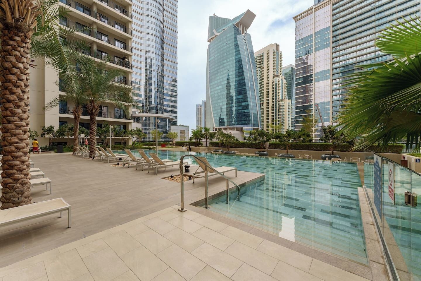 A rooftop pool with lounge chairs, palm trees, and modern high-rise buildings in the background.