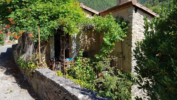 Un edificio de piedra rústica con jardín, muro de piedra y montañas al fondo.