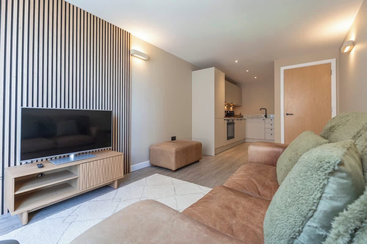 A modern living room with a flat-screen TV, a wooden entertainment unit, a sofa, and a kitchen in the background.
