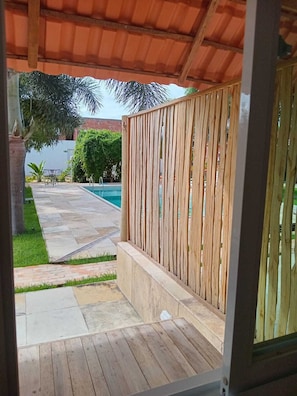 A wooden deck with a pool, a covered patio, and a view of a green lawn and trees.
