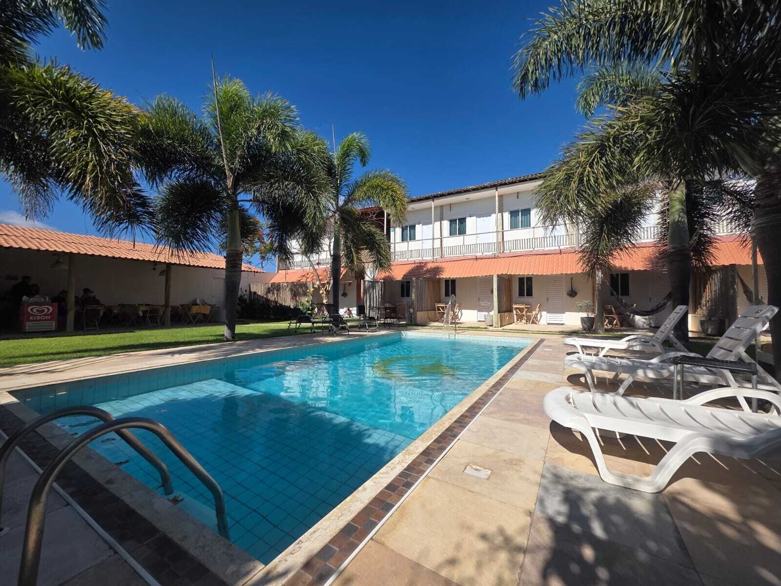 Piscina com espreguiçadeiras, palmeiras e um edifício com telhado vermelho.