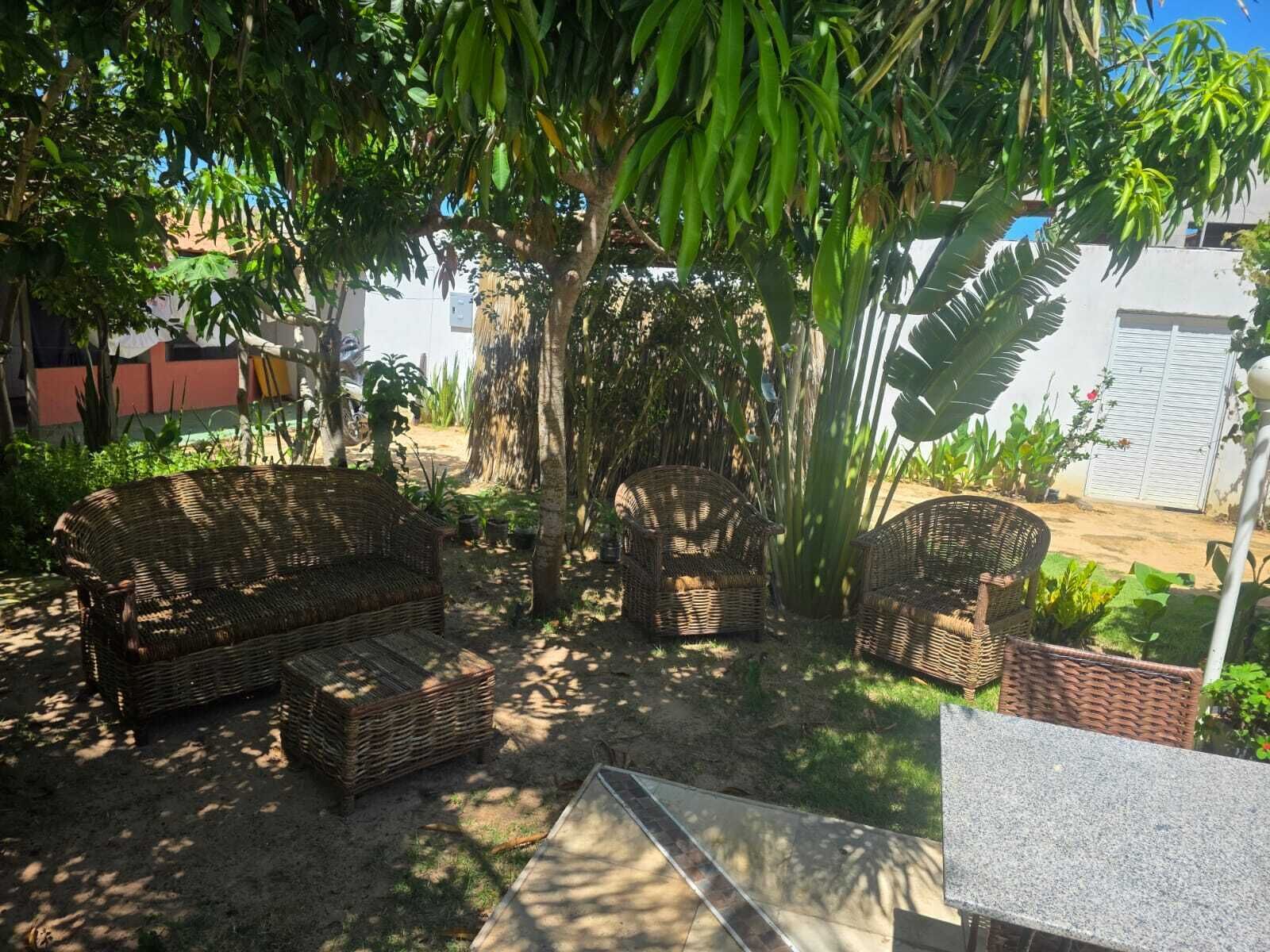 Un área de estar al aire libre con muebles de mimbre, una mesa, abundante vegetación y un edificio blanco al fondo.
