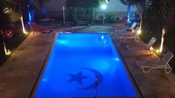 Una piscina dalla forma a stella, circondata da lettini e palme, illuminata di notte.