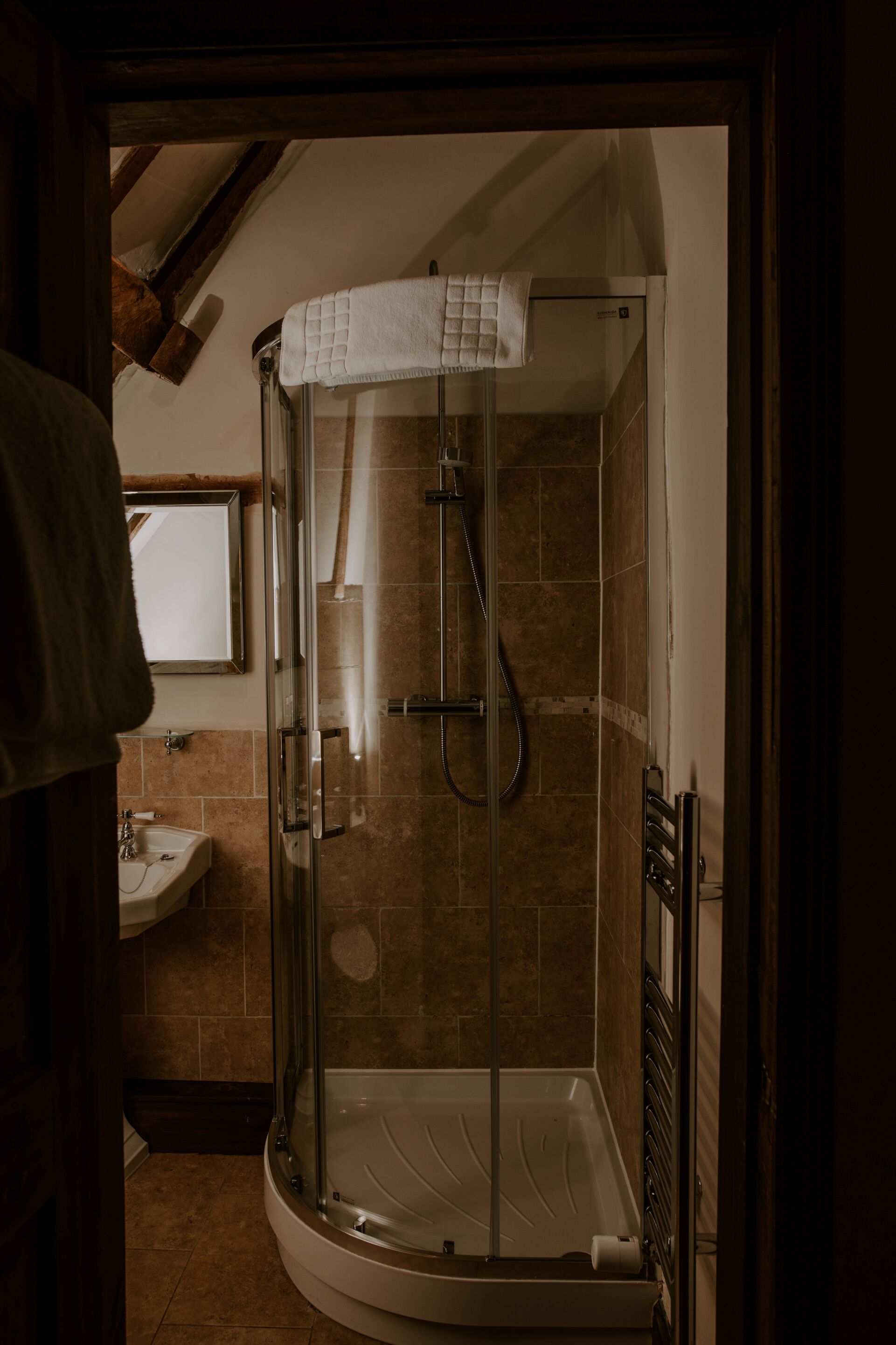 A glass-enclosed shower with a white towel, a sink, and a mirror in a bathroom with brown tiles.