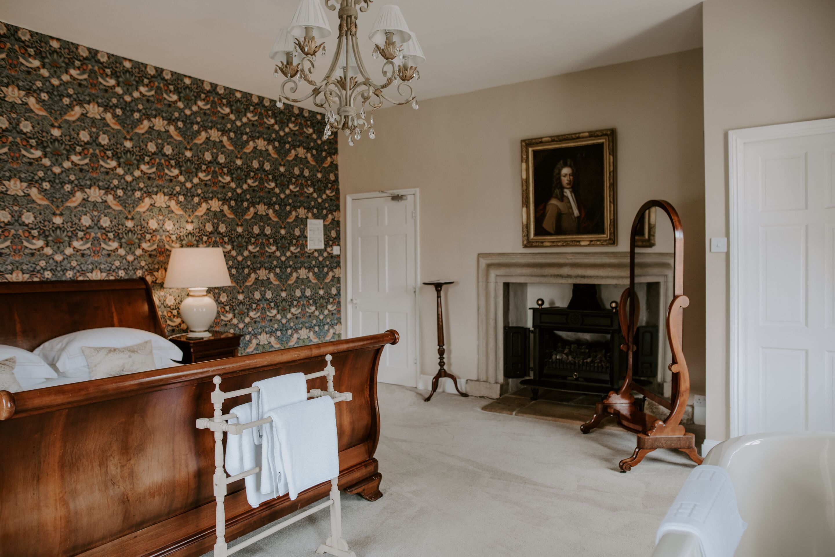 A bedroom with a wooden bed, a white bedside table, a chandelier, a fireplace, and a portrait on the wall.