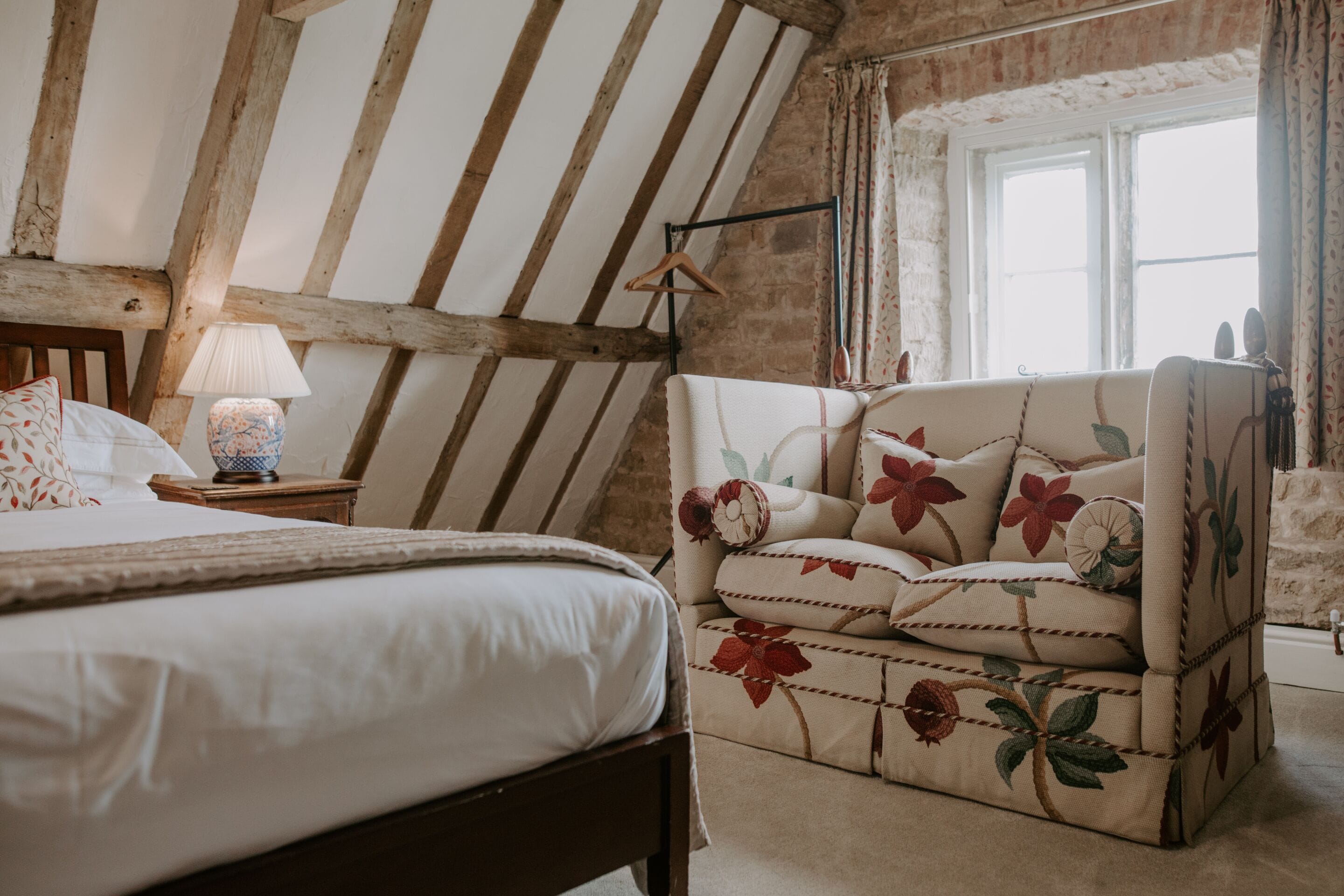 Un dormitorio con cama, una mesita de noche con lámpara, un sillón floral y una ventana con cortinas.