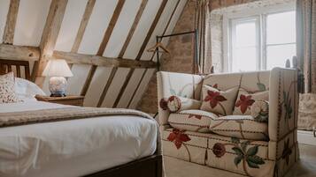 Un dormitorio con cama, una mesita de noche con lámpara, un sillón floral y una ventana con cortinas.