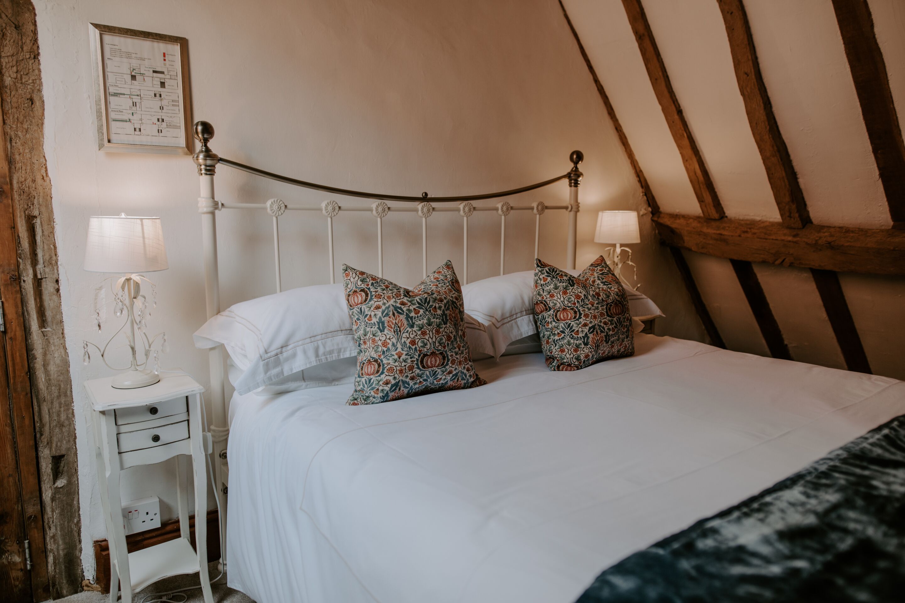 Un lit bien fait, agrémenté d’oreillers à motifs, une table de chevet blanche avec une lampe, et une poutre en bois au plafond.