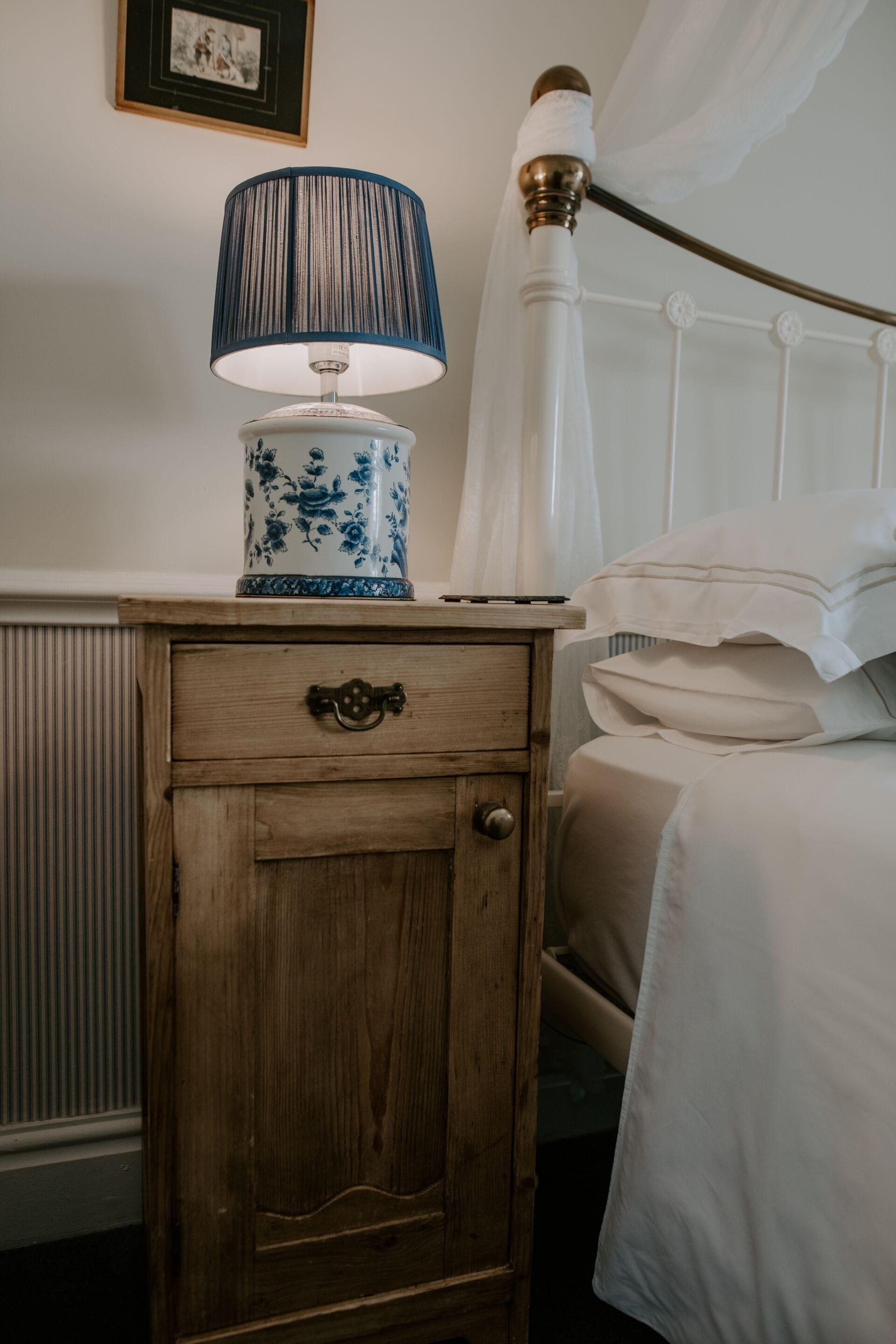 A wooden nightstand with a lamp and a framed picture on the wall above.