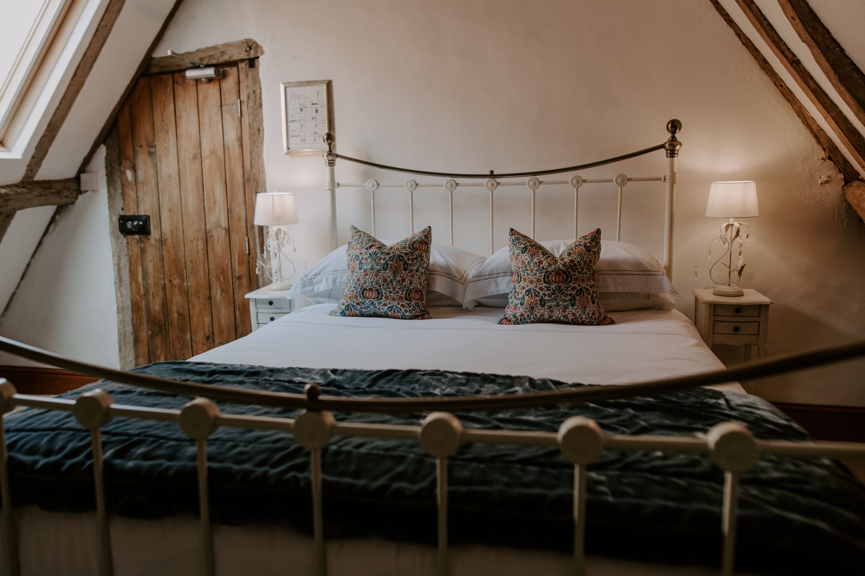 A bedroom with a sloped ceiling, a bed with patterned pillows, two bedside tables with lamps, and a wooden door.