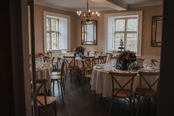 A dining room with multiple tables set for a meal, wooden chairs, a chandelier, and large windows with white shutters. - Allington Manor (Allington)
