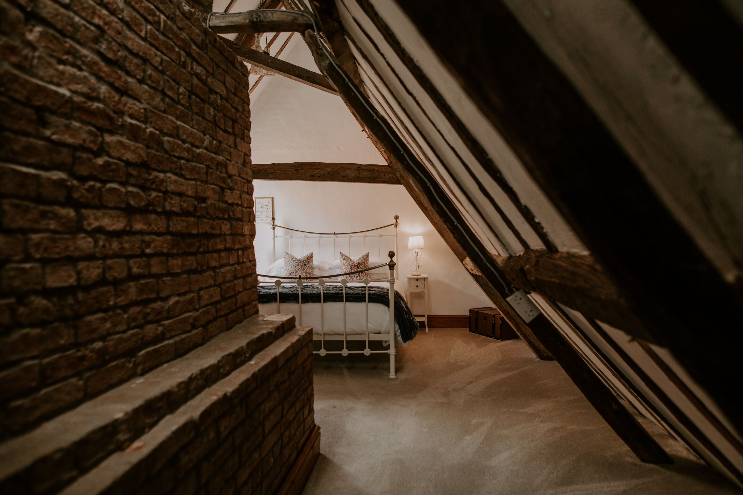 A bedroom with a sloped ceiling, a brick wall, a bed with a white frame, and a small bedside table with a lamp.