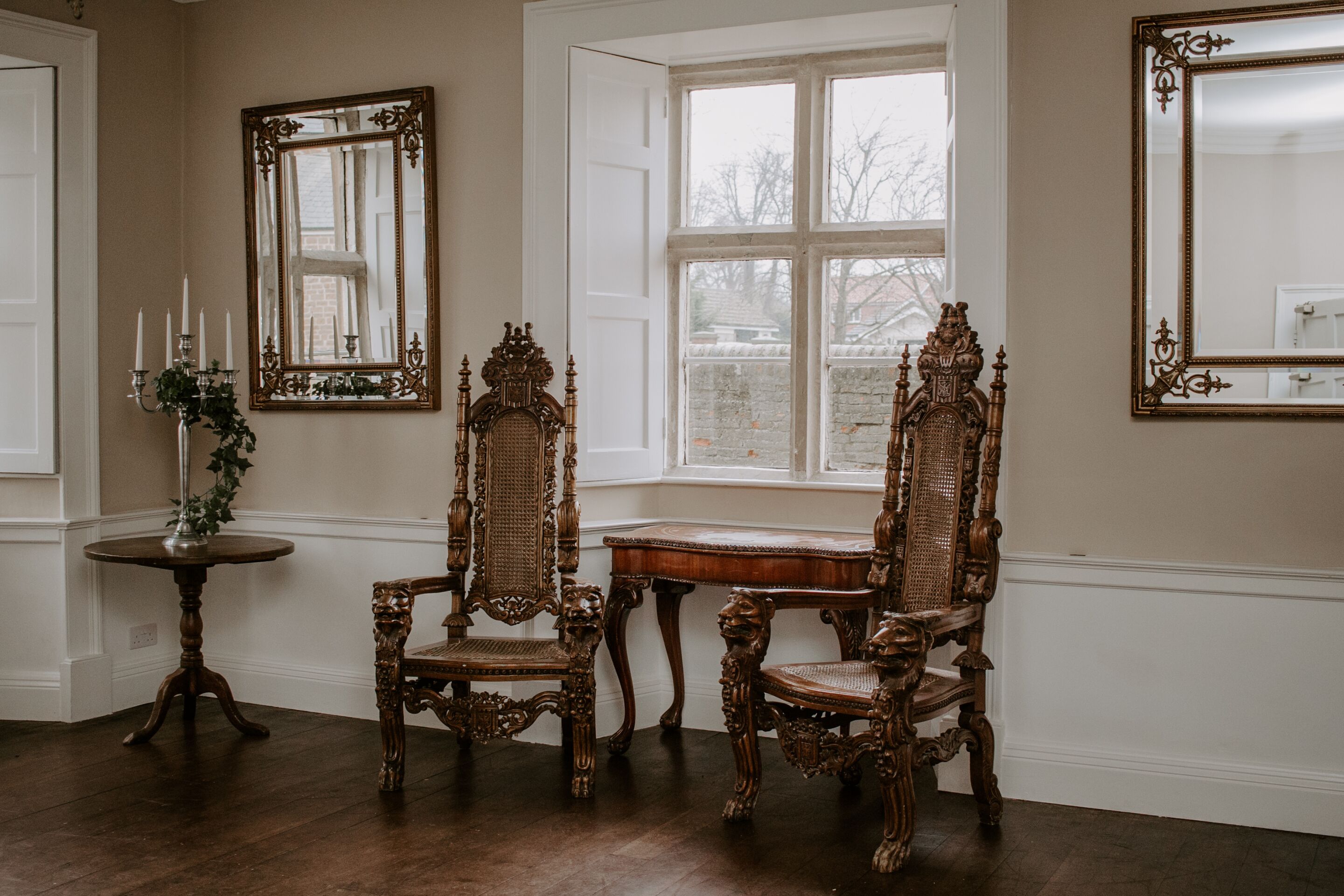 A room with ornate wooden furniture, including chairs and a table, positioned near a window with a view of the outside.