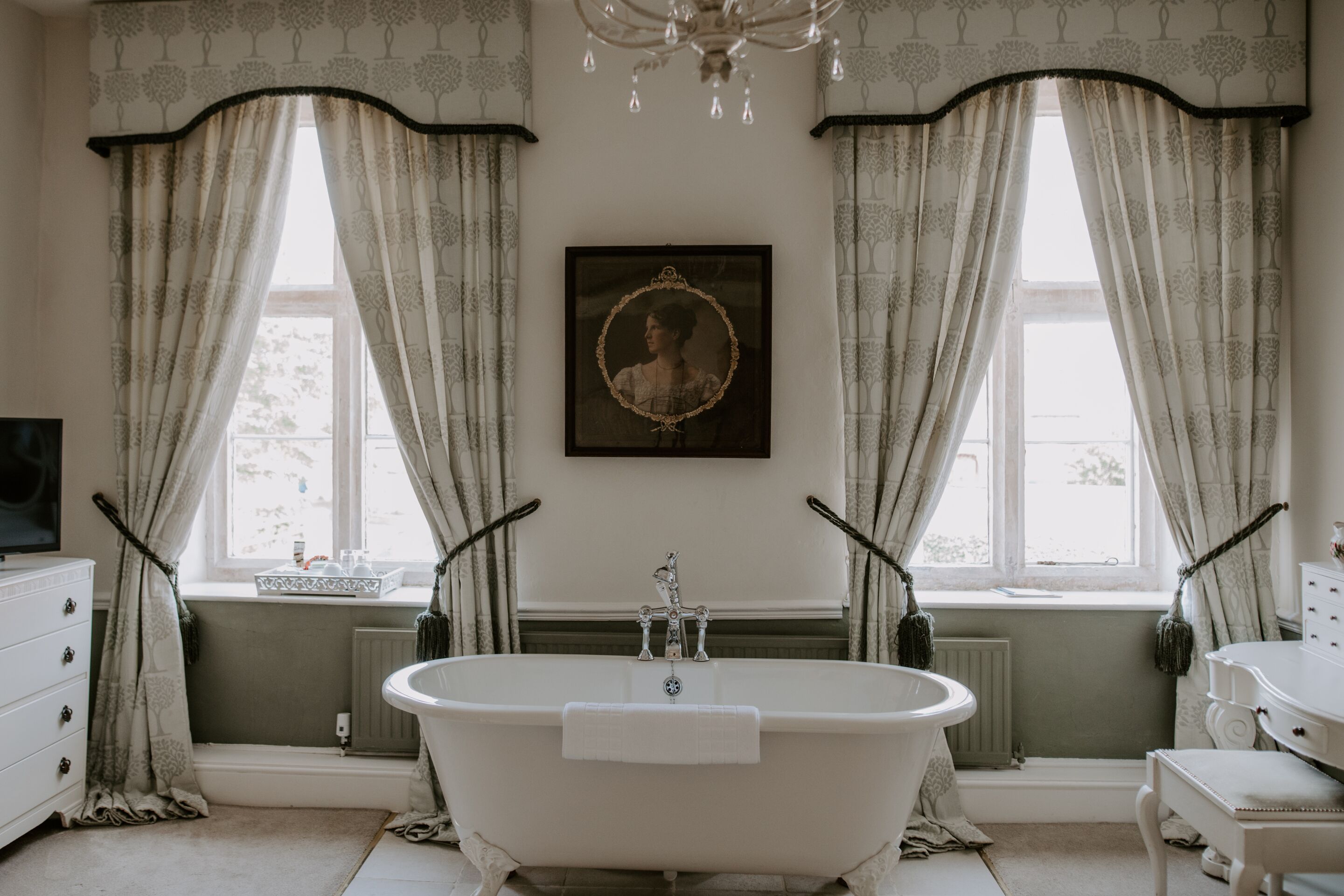 A bathroom with a freestanding bathtub, a radiator, a framed portrait, and a window with curtains.