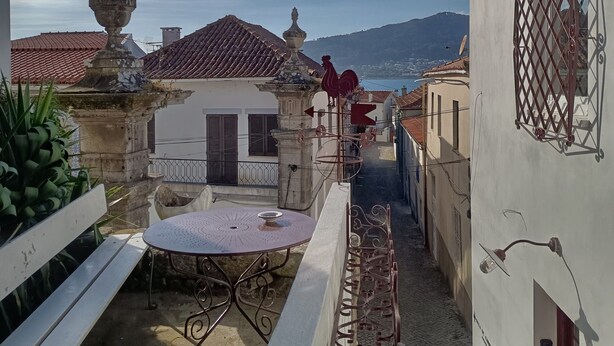 A courtyard with a white table, a red rooster statue, and a view of buildings with red roofs. - Portas de Caminha III Studio with Shared Terrace, Balcony and Wi-Fi (Caminha)