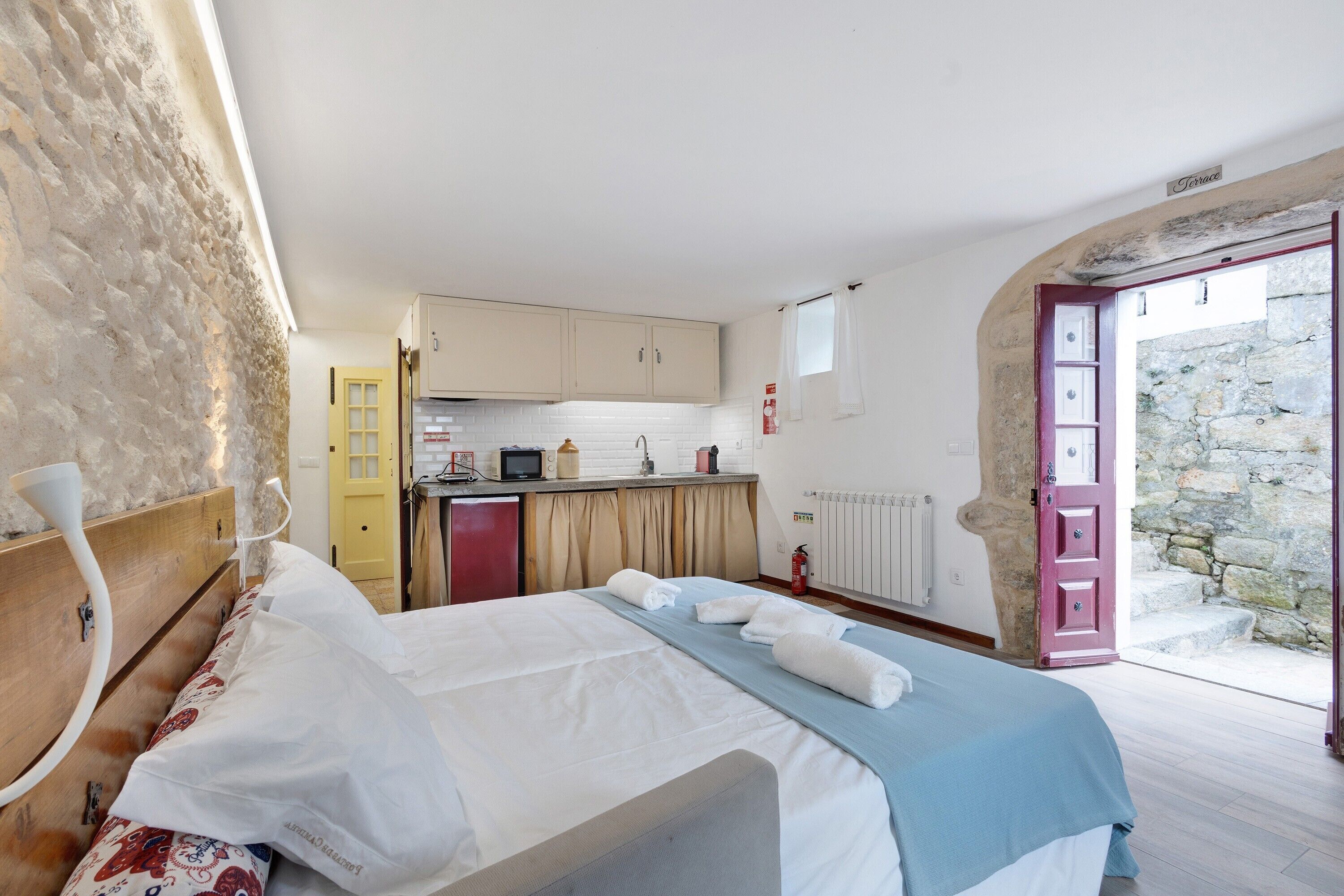 A bedroom with a bed, a small kitchenette, a stone wall, and a door leading to a balcony.