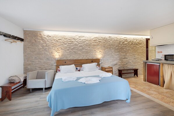 Un dormitorio con pared de piedra, cama con ropa blanca, cabecero de madera, mesita de noche y una pequeña kitchenette.