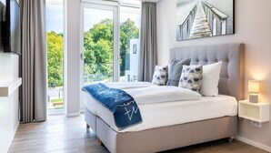Quarto moderno com cama grande, televisão, quadro e vista para o verde lá fora.