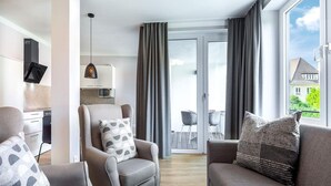 Uma sala moderna com sofá, poltrona e mesa de centro. Há uma cozinha com micro-ondas e armários, e uma varanda com vista.