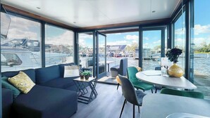A modern interior with a sofa, chairs, a table, and large windows offering a view of the water and boats. - Houseboat for 4 on Uitgeester Lake (Uitgeest)