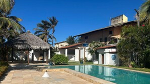 A pool area with a thatched-roof structure, a brick building, and palm trees. - Prainha - Aquiraz 5-bedroom summer mansion (Aquiraz)