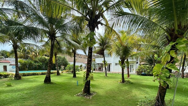 A well-maintained garden with palm trees, a swimming pool, and a white building with a red roof. - Prainha - Aquiraz 5-bedroom summer mansion (Aquiraz)