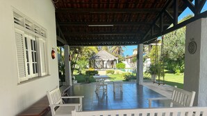 A covered patio with white benches, a thatched-roof structure, and a view of a garden and pool. - Prainha - Aquiraz 5-bedroom summer mansion (Aquiraz)