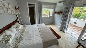 A bedroom with a bed, a bedside table, a mirror, a window with curtains, an air conditioning unit, and a balcony with a view of greenery. - Prainha - Aquiraz 5-bedroom summer mansion (Aquiraz)