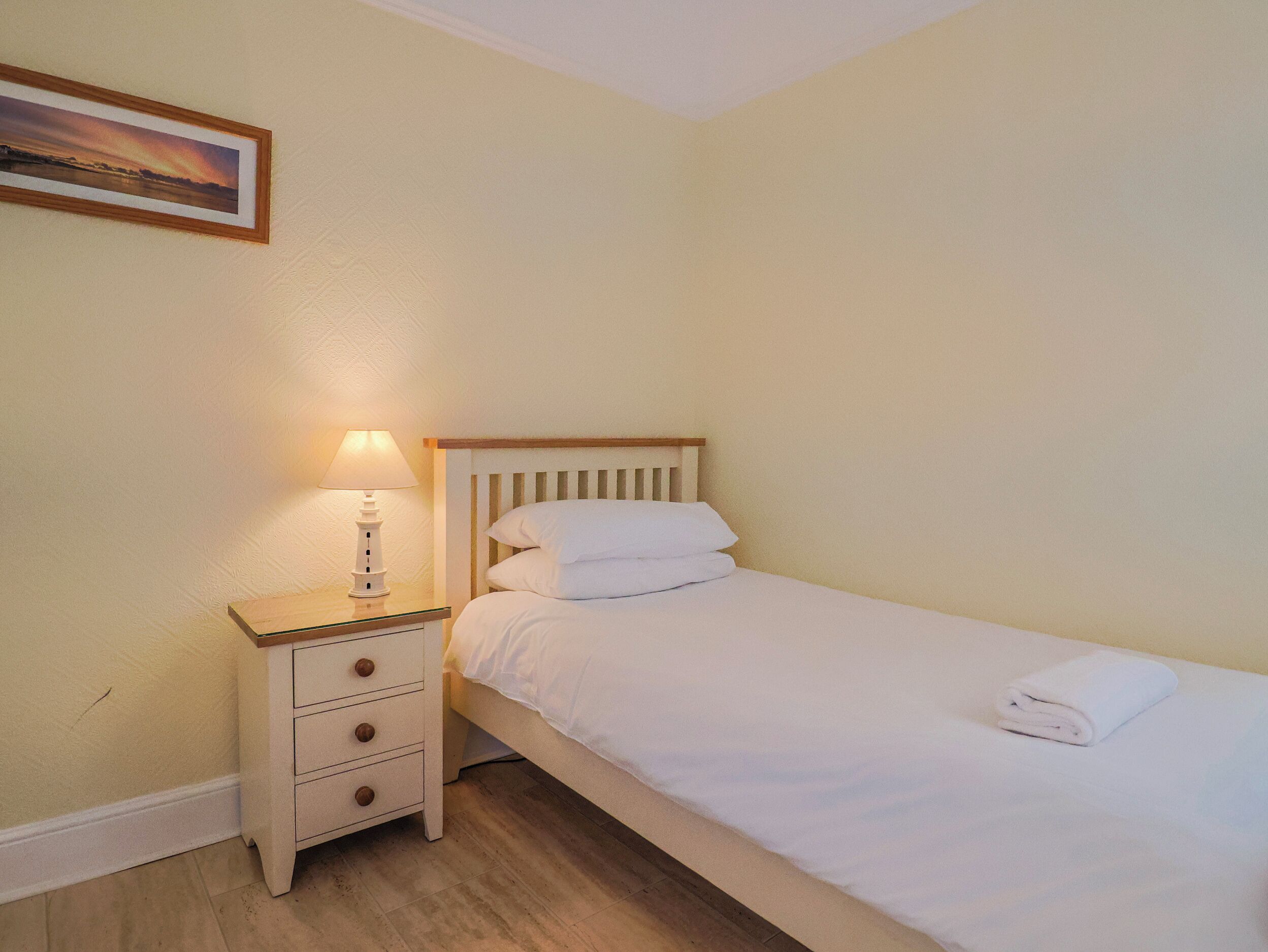 Un lit simple avec une tête de lit en bois, une table de chevet avec une lampe et un tableau encadré au mur.