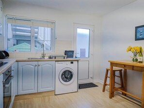 A compact kitchen with a washing machine, a sink, a window, and a wooden table with a vase of flowers. - Sea Lovers (Exmouth)