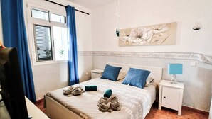 Un dormitorio con cama, dos almohadas, una mesita de noche con lámpara, una ventana con cortinas y un cuadro floral en la pared.
