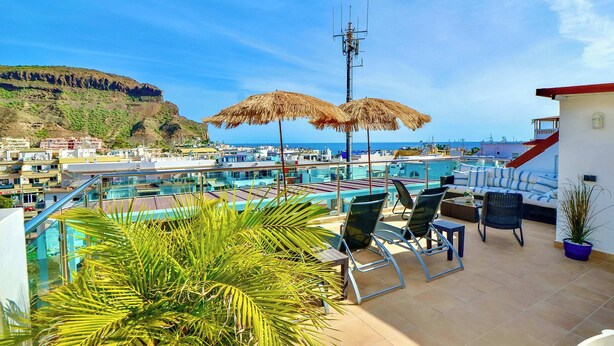 Una terraza en la azotea con piscina, sillas, sombrillas y vistas a la costa.