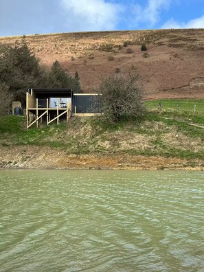 A riverside cabin with a deck and a small building, surrounded by a natural landscape. - Mountain Hidaway next to a Pool (Talerddig)