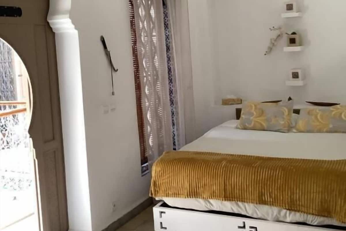 A bedroom with a bed, a hanging light fixture, a patterned curtain, and a small shelf with decorative items.