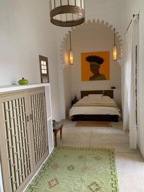 A narrow hallway leading to a bed with a patterned rug, a green bowl on a cabinet, and a painting on the wall.