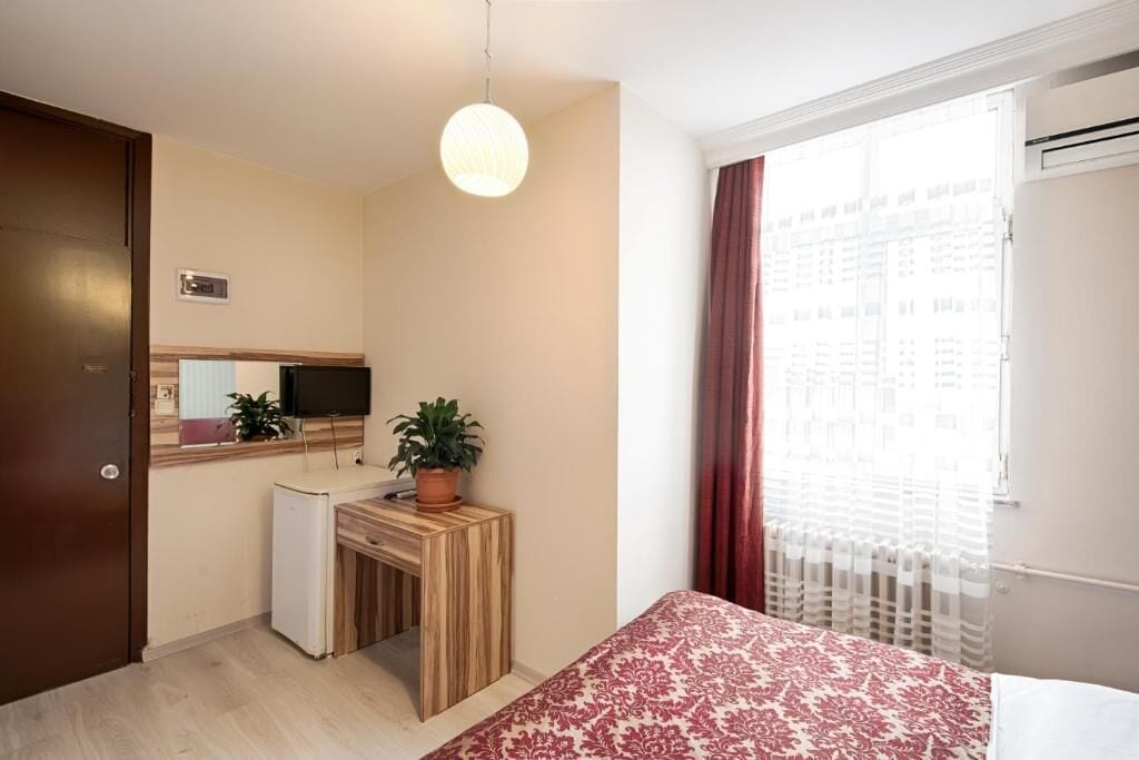 Ein Schlafzimmer mit einem Bett, einem Schreibtisch mit Pflanze, einem Fernseher und einem Fenster mit Vorhängen.