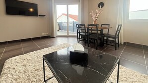 A modern living room with a flat-screen TV, a dining table with chairs, a coffee table with a tissue box, and a sliding glass door leading to a balcony. - Modern house with terrace and parking. Overlooking the city (Clermont-Ferrand)