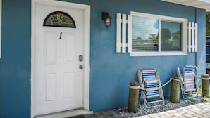 Una casa con puerta blanca, ventana con contraventanas blancas y dos sillas de jardín a rayas.