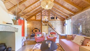 A rustic interior with a stone wall, wooden ceiling beams, a fireplace, and a cozy seating area with a sofa and two pink armchairs. - Holiday Home 'Eclectic Monte Laginha Alentejo' with Private Pool, Terrace & Wi-Fi (Lajinha)