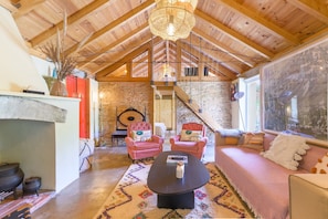 A rustic interior with a stone wall, wooden ceiling beams, a fireplace, and a cozy seating area with a sofa and two pink armchairs.
