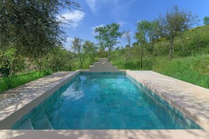 A rectangular swimming pool with a geometric structure in the center, surrounded by lush greenery and trees.