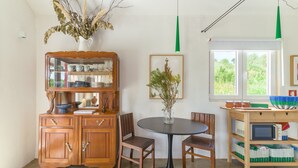 Een eetruimte met een houten kast, een ronde tafel, stoelen, een vaas met gedroogde planten, een ingelijst beeld en een raam met uitzicht op groen.