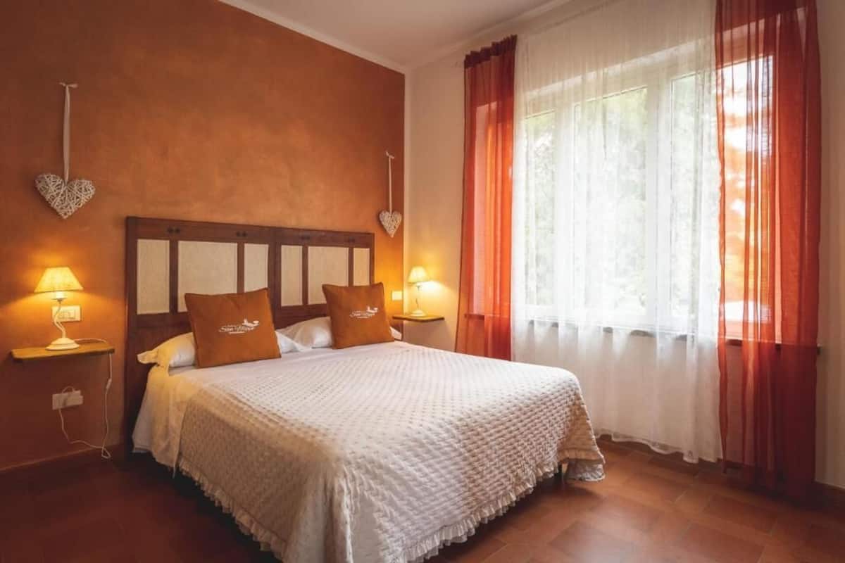 A hotel room with a wooden headboard, two bedside lamps, a bed with white bedding, and a window with orange curtains.