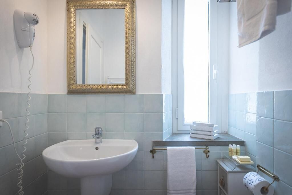 Ein Badezimmer mit einem weißen Waschbecken, einem spiegel mit goldenem Rahmen, einem Handtuchhalter und einem Fenster.