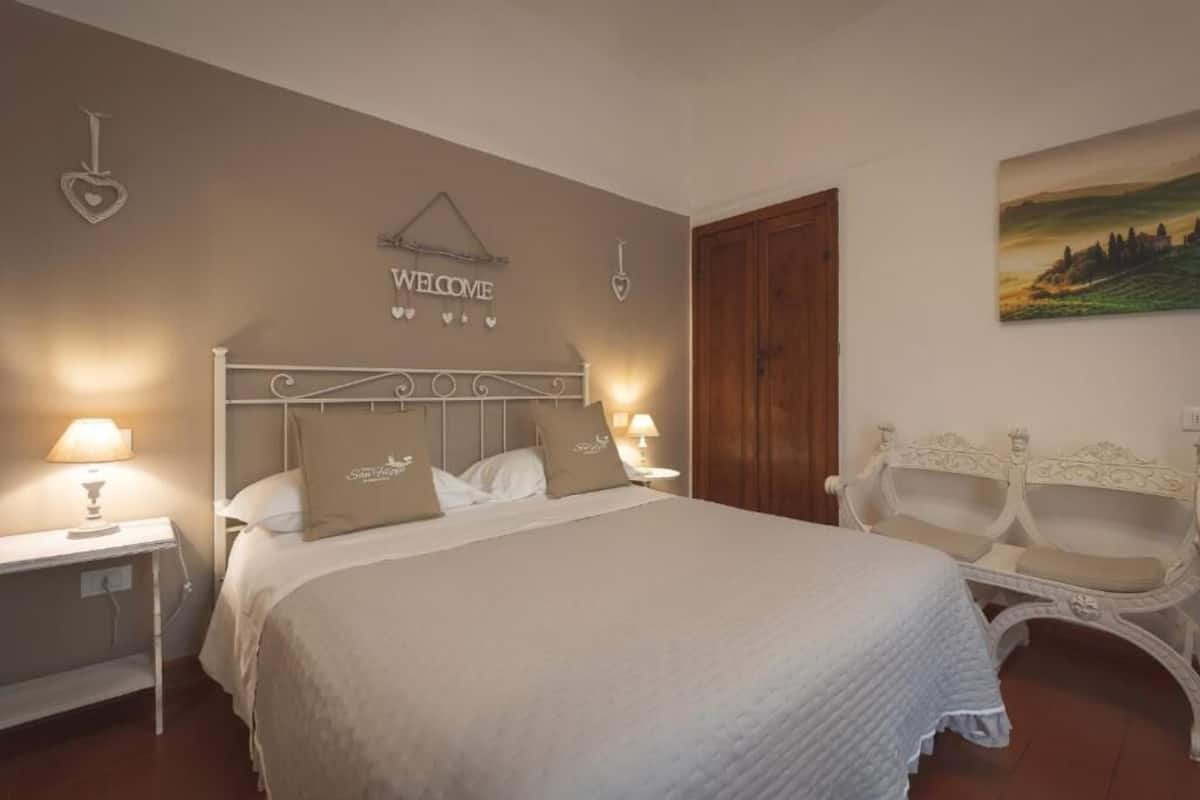A bedroom with a bed, bedside tables, a bench, a 'WELCOME' sign, and a landscape painting.