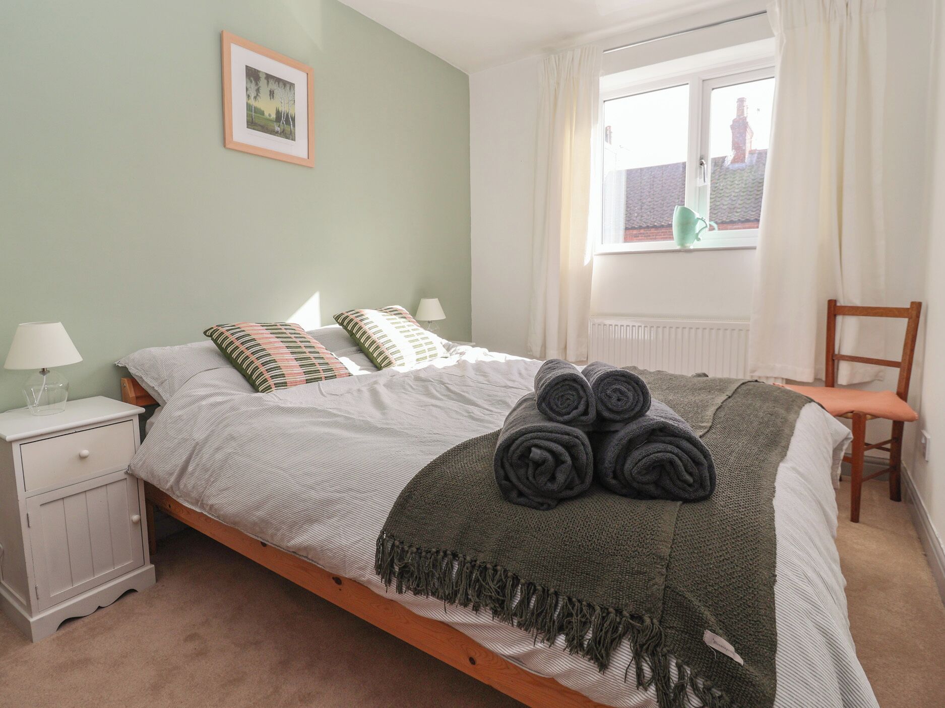 A bedroom with a bed, a nightstand, a chair, a window with curtains, and a framed picture on the wall.